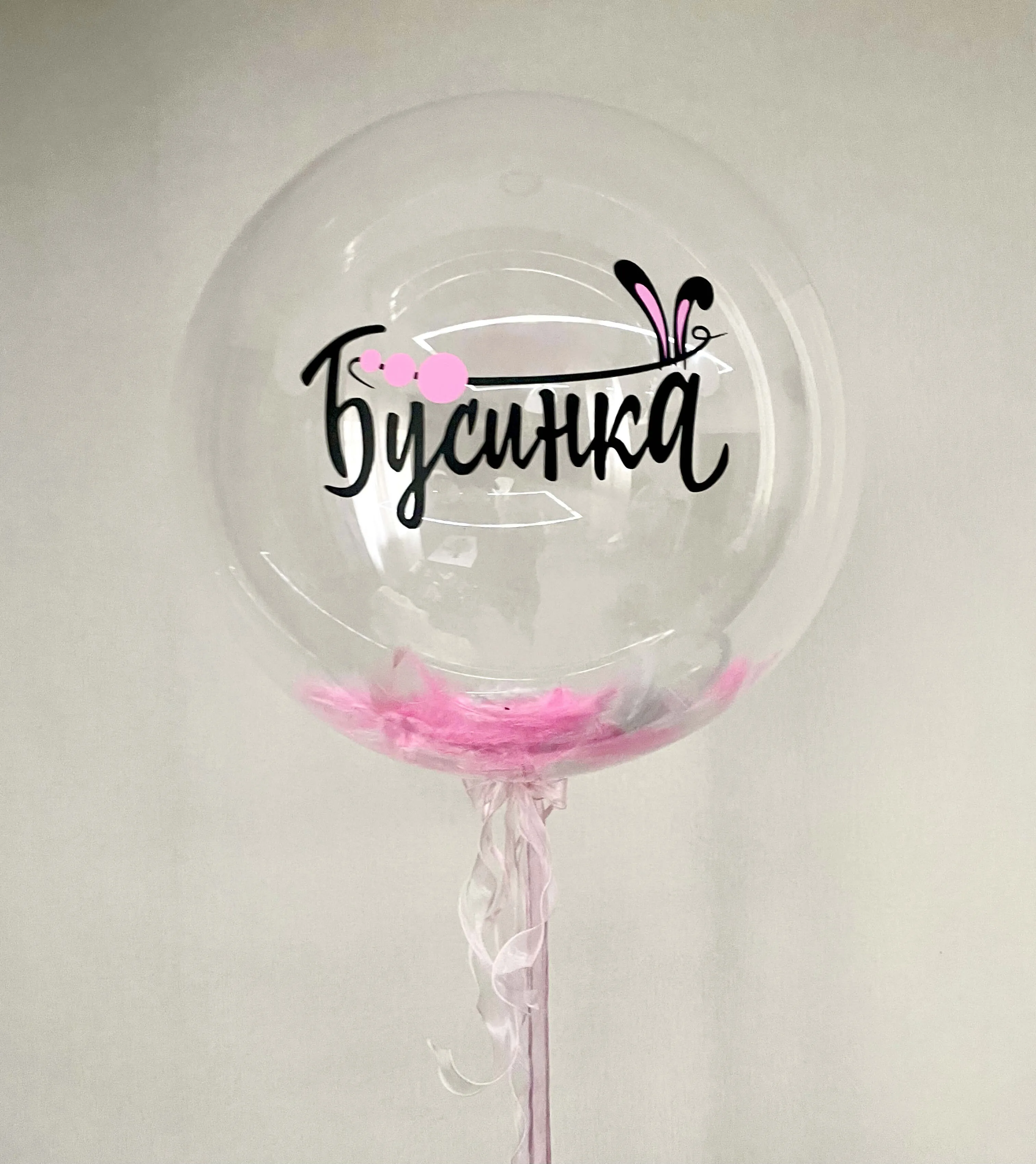 Декоративный прозрачный шар с именем „Бусинка“, написанным чёрным курсивом, с розовыми перьями в нижней части и лентой для крепления